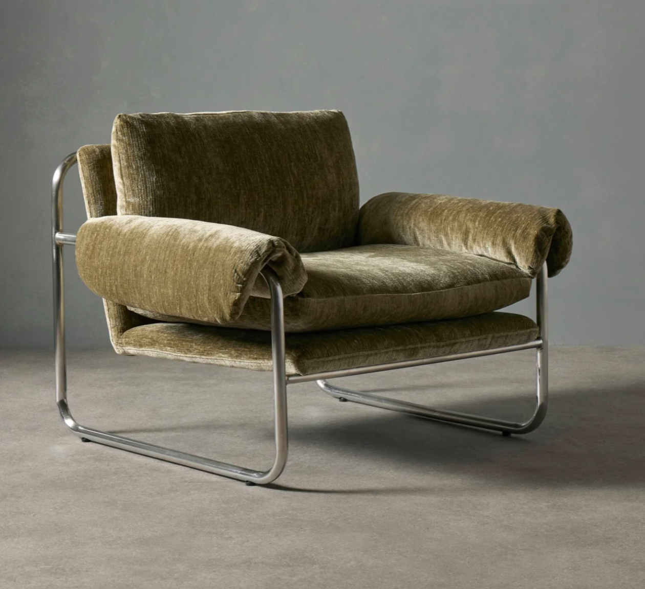 A mid-century modern armchair with plush olive green velvet upholstery and a curved chrome metal frame, set against a neutral gray studio background.