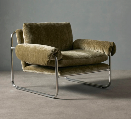 A mid-century modern armchair with plush olive green velvet upholstery and a curved chrome metal frame, set against a neutral gray studio background.